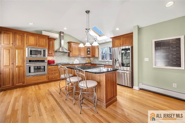 kitchen featuring wall chimney range hood, brown cabinetry, stainless steel appliances, a kitchen island, and a baseboard heating unit