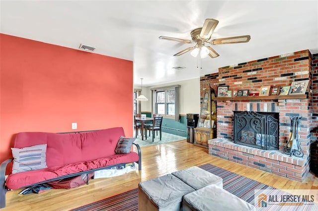 living area featuring a brick fireplace, ceiling fan, visible vents, and wood finished floors