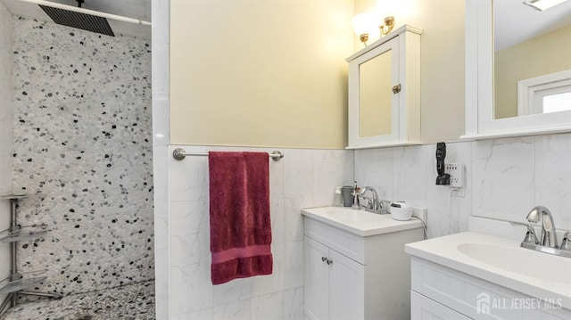 bathroom with tile walls, tiled shower, two vanities, and a sink