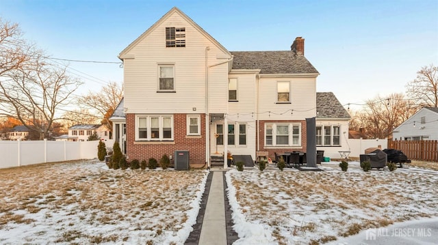 snow covered house featuring brick siding, entry steps, central AC unit, a chimney, and a fenced backyard