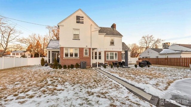 snow covered back of property with cooling unit, a fenced backyard, a chimney, entry steps, and brick siding