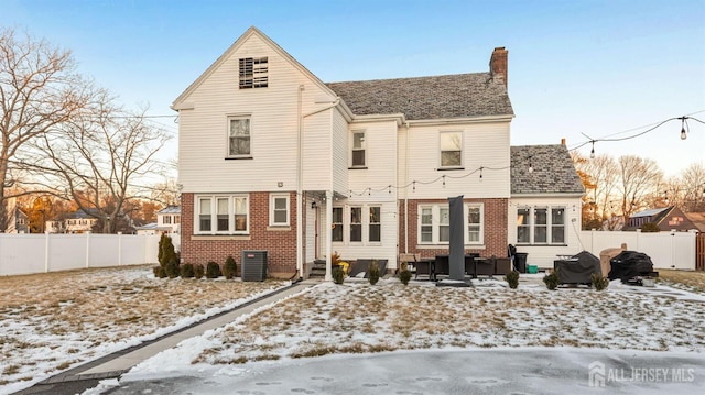 snow covered back of property featuring cooling unit, fence, an outdoor living space, a chimney, and brick siding