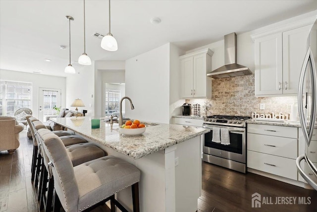 kitchen with white cabinets, wall chimney range hood, open floor plan, appliances with stainless steel finishes, and light stone countertops
