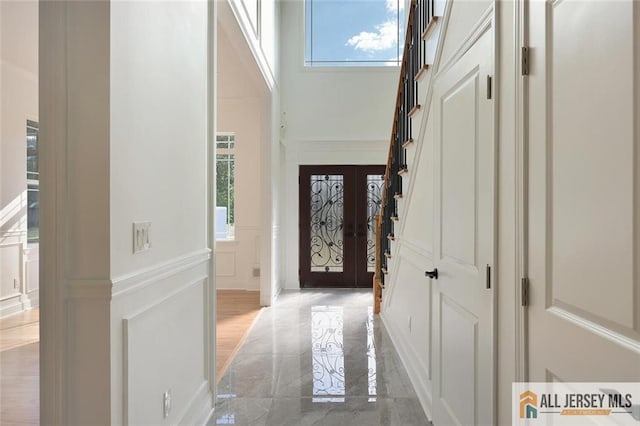 foyer entrance with a decorative wall, a wainscoted wall, a towering ceiling, marble finish floor, and french doors