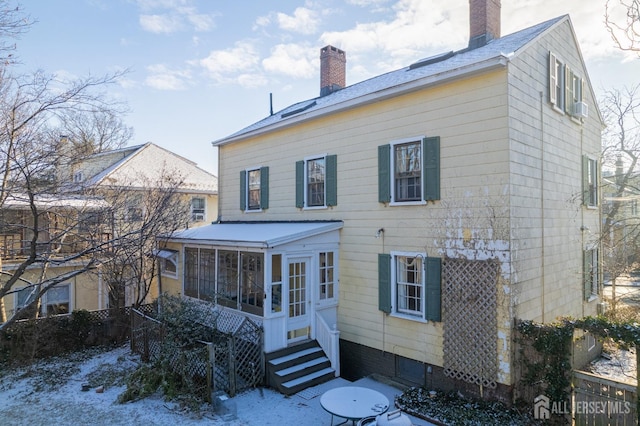 snow covered back of property with entry steps, a chimney, and a sunroom