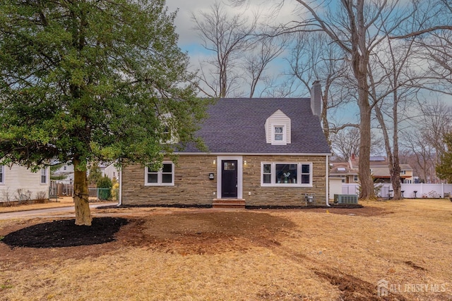 new england style home featuring cooling unit, a shingled roof, fence, stone siding, and a chimney