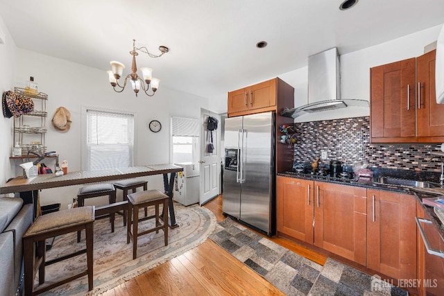 kitchen featuring a sink, decorative backsplash, wall chimney exhaust hood, brown cabinetry, and stainless steel fridge