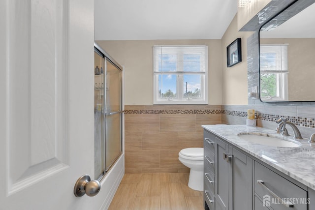 bathroom featuring vanity, toilet, enclosed tub / shower combo, a wainscoted wall, and tile walls