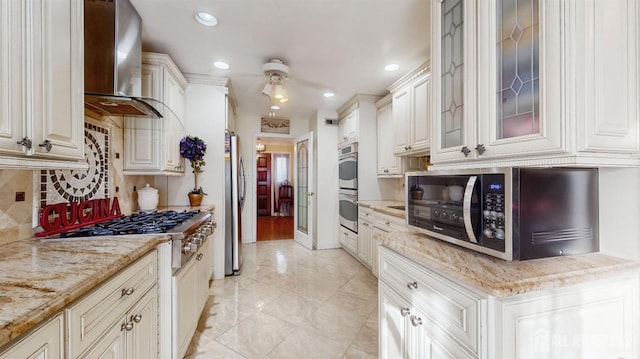 kitchen featuring white cabinetry, stainless steel appliances, light stone countertops, backsplash, and wall chimney range hood