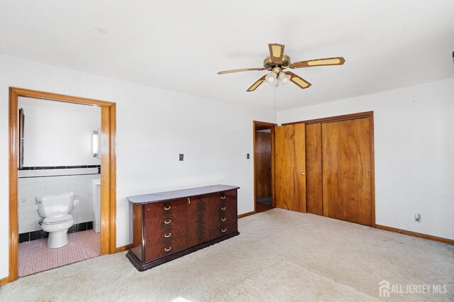 unfurnished bedroom featuring light carpet, ensuite bath, ceiling fan, tile walls, and a closet