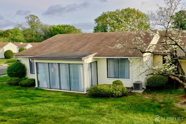 rear view of property featuring a lawn, cooling unit, a shingled roof, and stucco siding