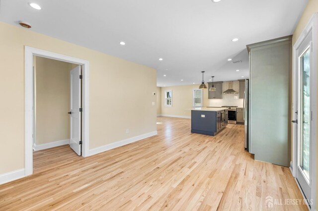 kitchen with appliances with stainless steel finishes, pendant lighting, a kitchen island, wall chimney exhaust hood, and light hardwood / wood-style flooring