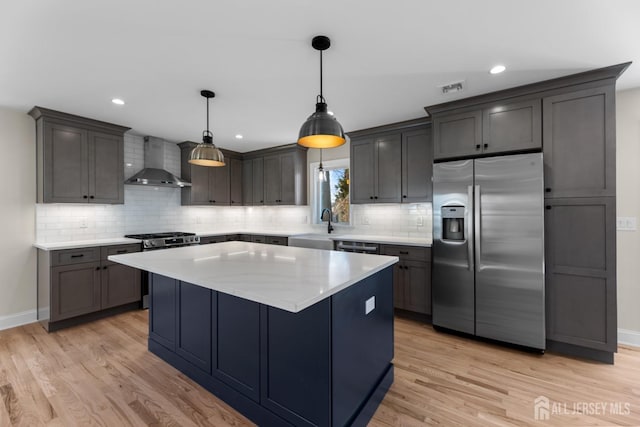 kitchen with appliances with stainless steel finishes, hanging light fixtures, light wood-type flooring, a center island, and wall chimney range hood