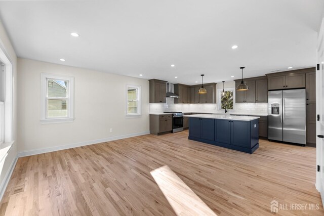 kitchen with decorative light fixtures, wall chimney range hood, a kitchen island, appliances with stainless steel finishes, and dark brown cabinets