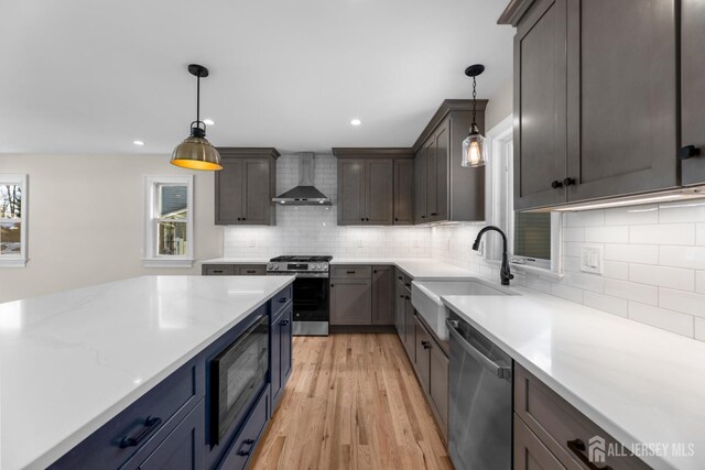 kitchen with light stone counters, wall chimney exhaust hood, light hardwood / wood-style flooring, pendant lighting, and appliances with stainless steel finishes