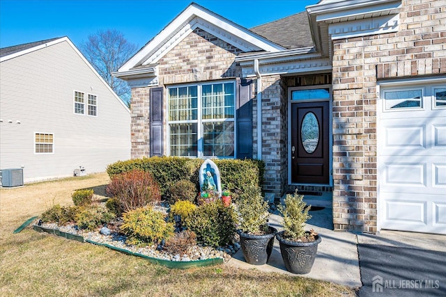 doorway to property featuring an attached garage, a shingled roof, central AC unit, and brick siding