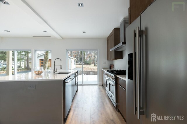 kitchen with a sink, high quality appliances, light countertops, wall chimney range hood, and a center island with sink