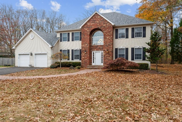 colonial house with aphalt driveway, roof with shingles, an attached garage, fence, and brick siding