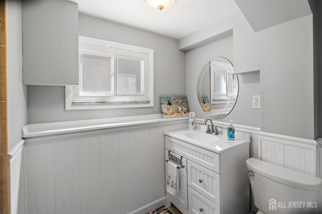 half bath with wainscoting, vanity, toilet, healthy amount of natural light, and a decorative wall