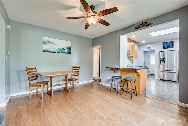 kitchen with stainless steel fridge with ice dispenser, a breakfast bar, a skylight, ceiling fan, and light wood-type flooring