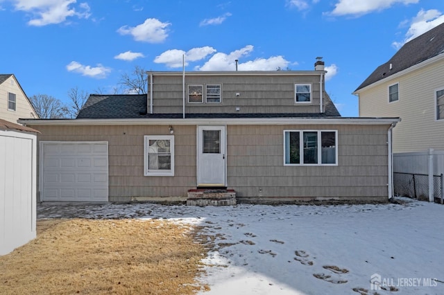 snow covered back of property featuring an attached garage, fence, and roof with shingles