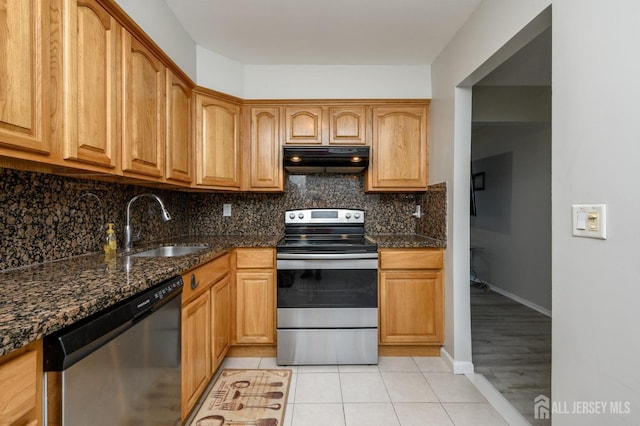 kitchen featuring light tile patterned floors, dark stone counters, appliances with stainless steel finishes, under cabinet range hood, and a sink