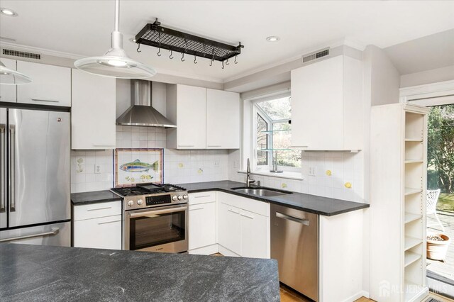 kitchen with decorative light fixtures, sink, white cabinets, stainless steel appliances, and wall chimney range hood