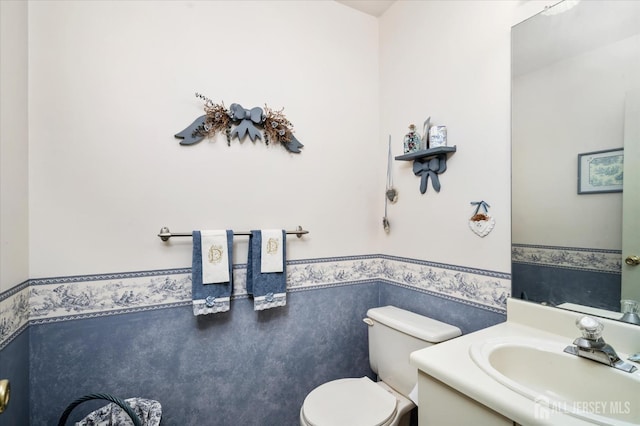 half bathroom featuring toilet, a wainscoted wall, wallpapered walls, and vanity