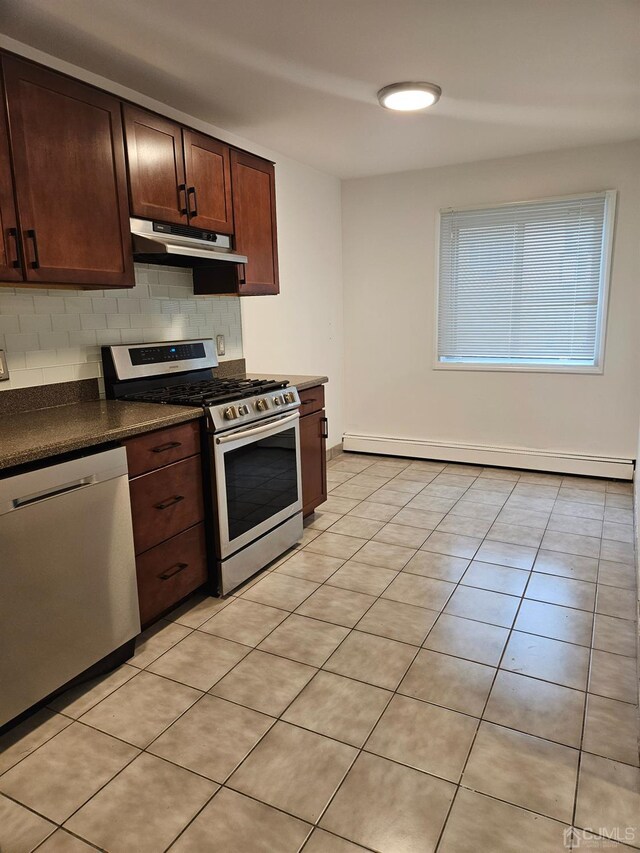 kitchen with dark countertops, backsplash, under cabinet range hood, baseboard heating, and appliances with stainless steel finishes