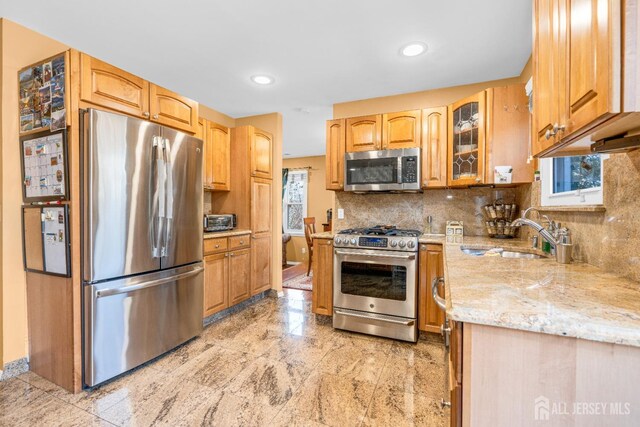 kitchen with light stone counters, a sink, stainless steel appliances, backsplash, and glass insert cabinets