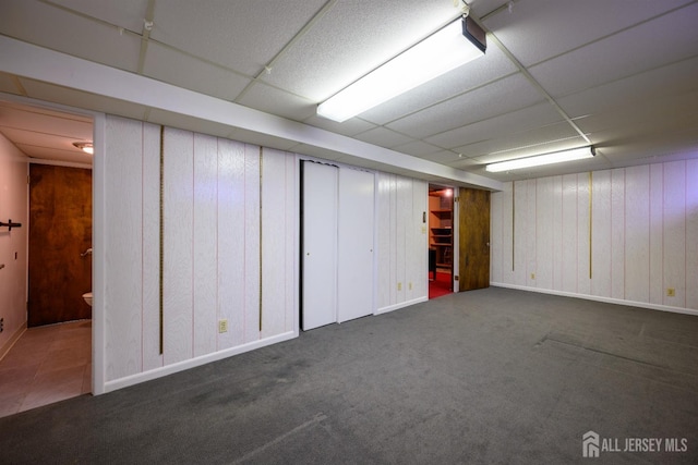 basement featuring a drop ceiling, wooden walls, and dark colored carpet