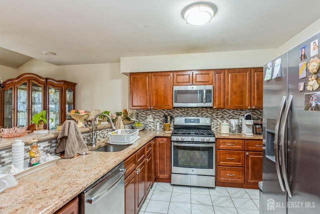 kitchen with light stone counters, stainless steel appliances, a sink, backsplash, and brown cabinets