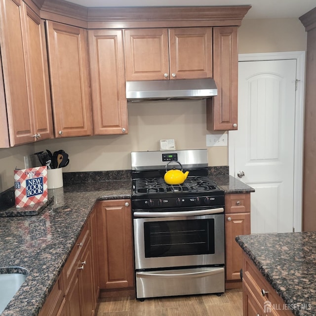 kitchen with brown cabinetry, stainless steel gas stove, under cabinet range hood, and dark stone countertops