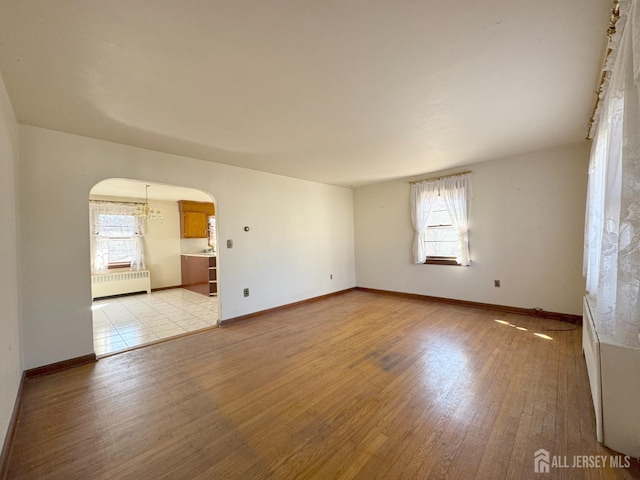 empty room featuring arched walkways, light wood-style flooring, radiator, and baseboards