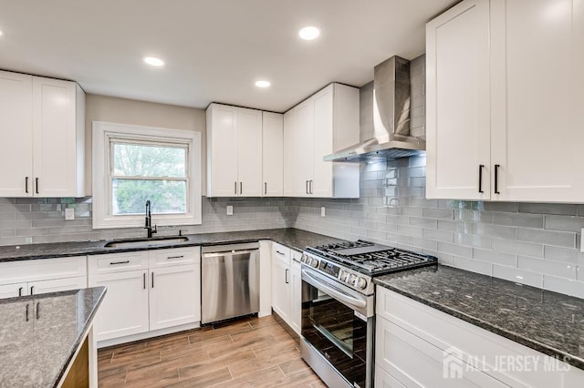 kitchen featuring stainless steel appliances, wall chimney range hood, a sink, dark stone countertops, and white cabinetry
