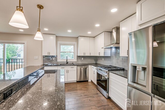 kitchen with appliances with stainless steel finishes, wall chimney exhaust hood, a sink, hanging light fixtures, and dark stone countertops