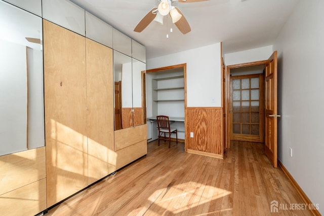 interior space with built in desk, a wainscoted wall, ceiling fan, and light wood-style floors