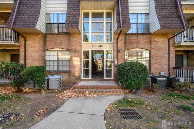 property entrance featuring central AC, mansard roof, brick siding, and roof with shingles