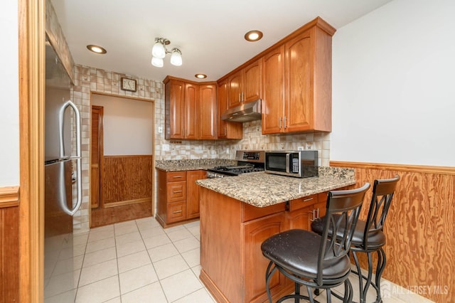 kitchen featuring a wainscoted wall, under cabinet range hood, stainless steel appliances, a peninsula, and light tile patterned flooring