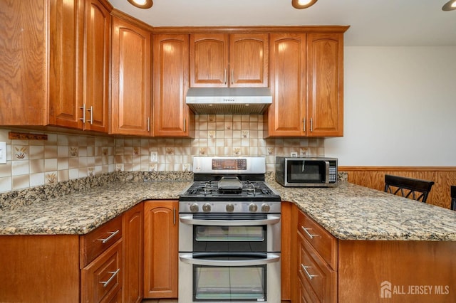 kitchen with under cabinet range hood, appliances with stainless steel finishes, brown cabinetry, and light stone counters