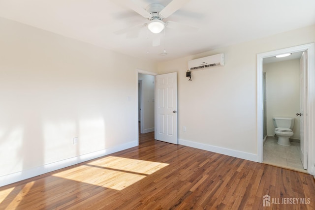 unfurnished bedroom with connected bathroom, baseboards, a wall mounted air conditioner, a ceiling fan, and wood-type flooring