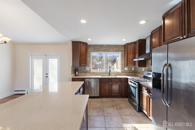 kitchen featuring a baseboard heating unit, wall chimney range hood, decorative backsplash, stainless steel appliances, and a sink