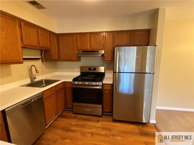 kitchen featuring stainless steel appliances, light countertops, brown cabinetry, a sink, and under cabinet range hood