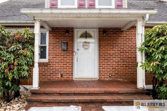 view of exterior entry featuring brick siding, covered porch, and roof with shingles