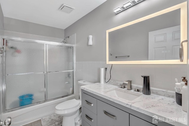 full bathroom featuring vanity, bath / shower combo with glass door, tile walls, and toilet