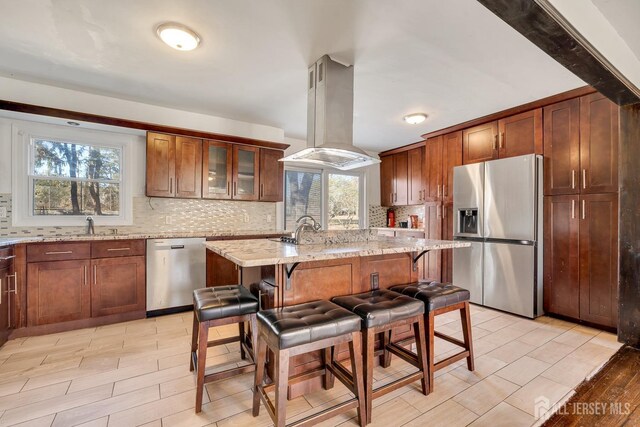 kitchen with a breakfast bar, a center island with sink, island exhaust hood, stainless steel appliances, and light stone countertops