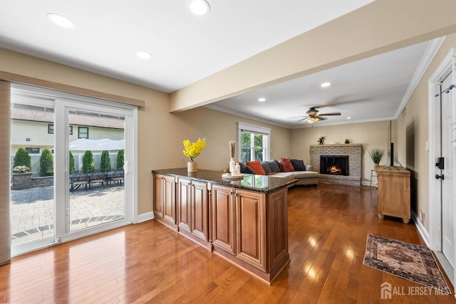 kitchen with a brick fireplace, dark stone countertops, ceiling fan, hardwood / wood-style flooring, and ornamental molding