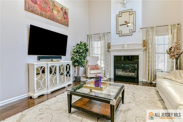 living room with baseboards, a fireplace with flush hearth, a towering ceiling, wood finished floors, and a ceiling fan