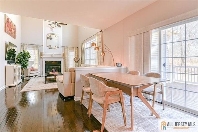 dining space with a glass covered fireplace, baseboards, a ceiling fan, and dark wood-style flooring