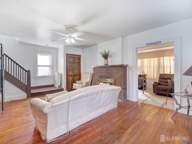 living room with wood finished floors, stairway, a brick fireplace, a ceiling fan, and baseboards
